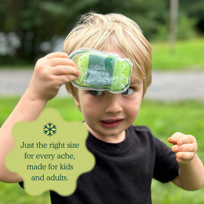 A young blonde boy outdoors holding a small green packaged heat pack against his forehead. The package has a clear window showing a cooling gel pad, and a pale green text bubble on the left reads, 'Just the right size for every ache, made for kids and adults.